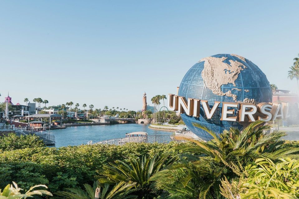 L'iconico logo blu del globo Universal si trova accanto a un corso d'acqua, con palme lussureggianti ed edifici sullo sfondo sotto un cielo sereno.