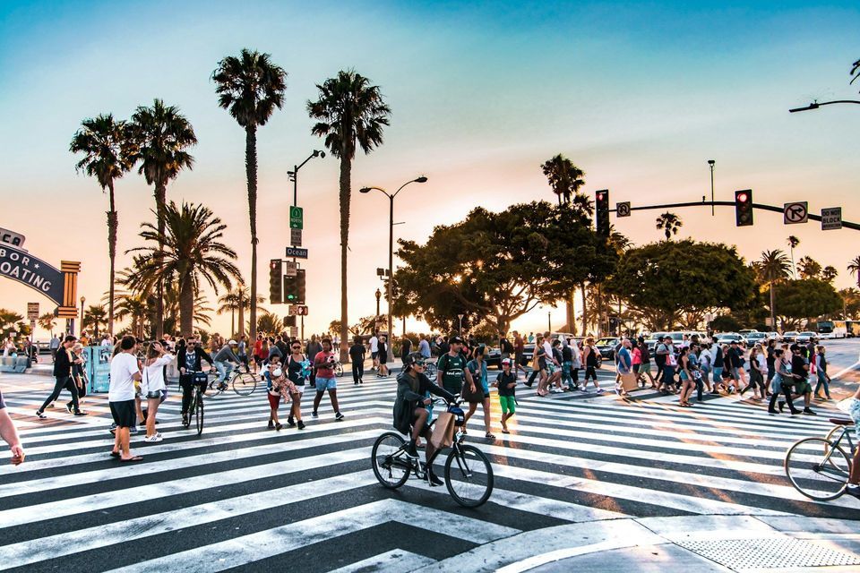 Un trafficato attraversamento pedonale con pedoni e ciclisti su una strada fiancheggiata da alte palme, sotto un cielo al tramonto.