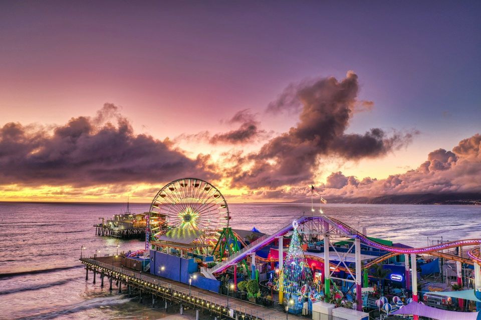 Un parco divertimenti illuminato su un molo sull'oceano, con una ruota panoramica e un albero di Natale sotto un cielo al tramonto colorato.