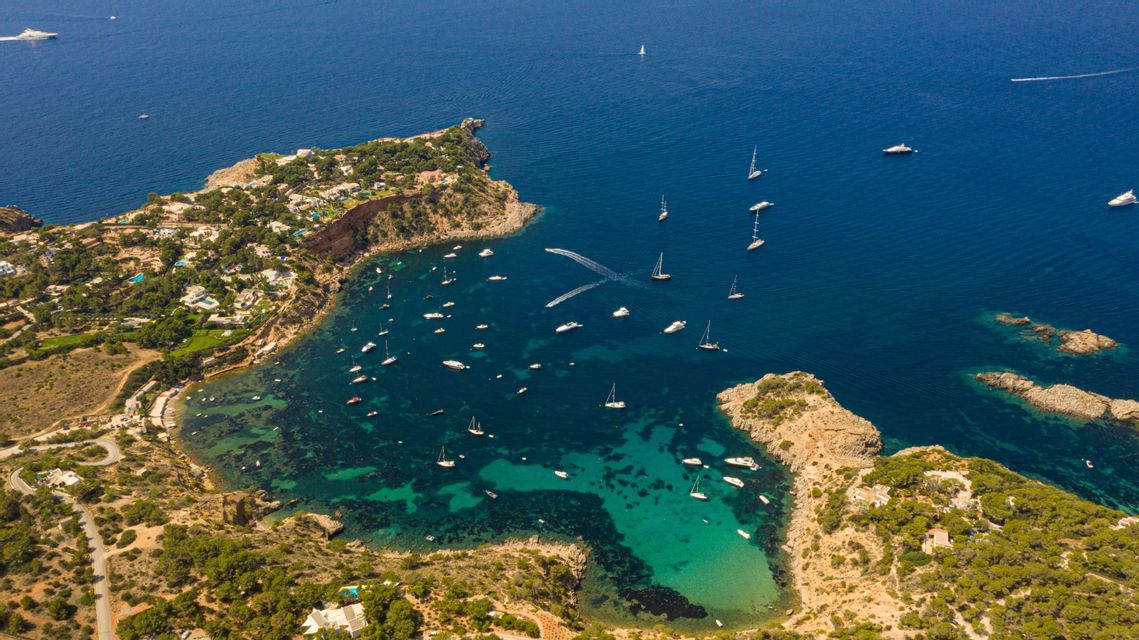 Vista aérea de numerosos barcos y yates anclados en una bahía tranquila y turquesa junto a una costa verde y rocosa.