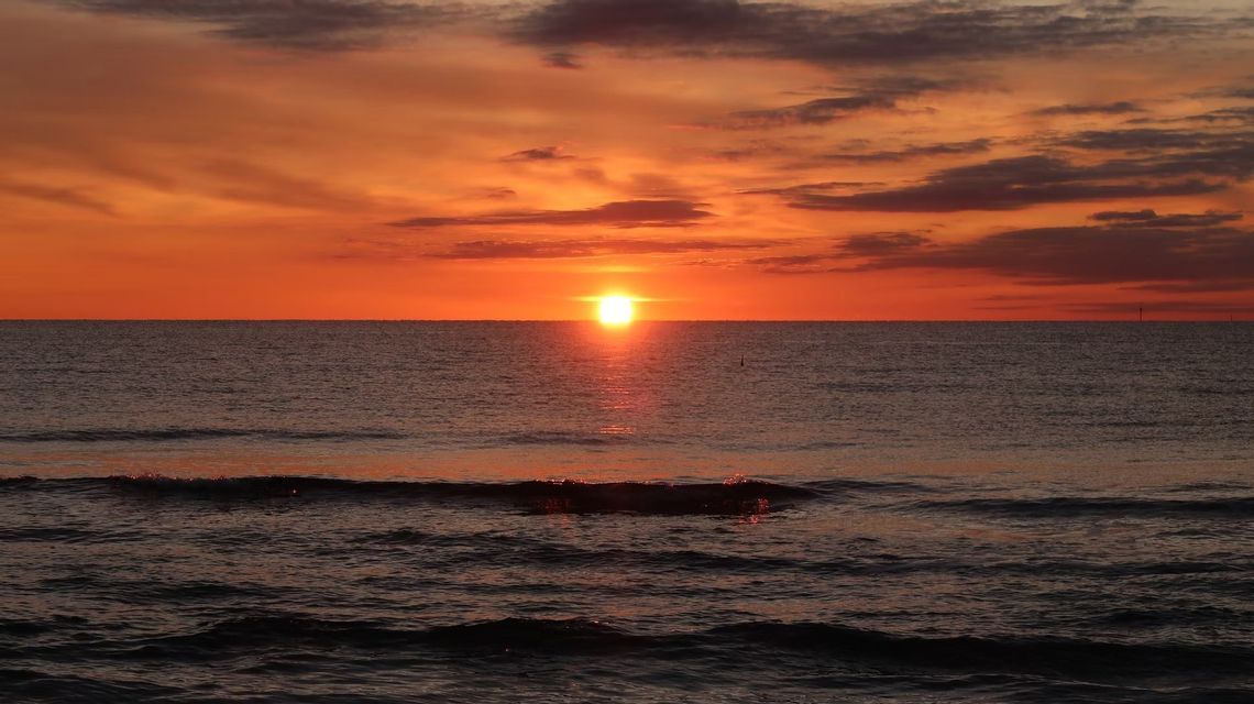 Le soleil se couche à l'horizon sur l'océan sous un ciel nuageux et orangé, avec sa lumière se reflétant sur l'eau.