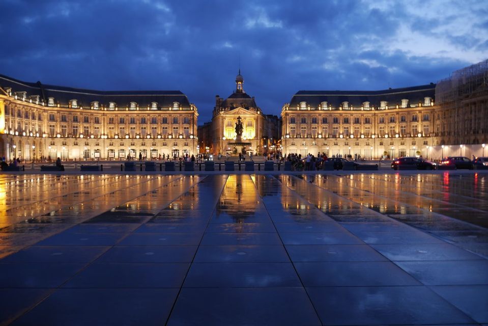 Des bâtiments classiques illuminés se reflètent sur la surface mouillée d'une grande place publique au crépuscule.