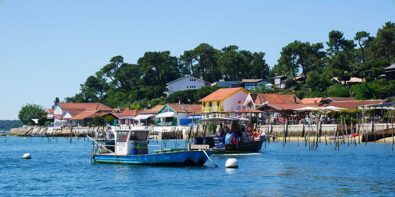 Deux bateaux sur une eau bleue calme devant un village au bord de l'eau avec des maisons colorées, des restaurants en plein air et des arbres verts.