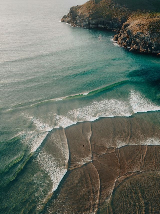 Vista aerea di surfisti in acque turchesi vicino a una spiaggia sabbiosa e a una scogliera rocciosa, con onde che si infrangono sulla riva.