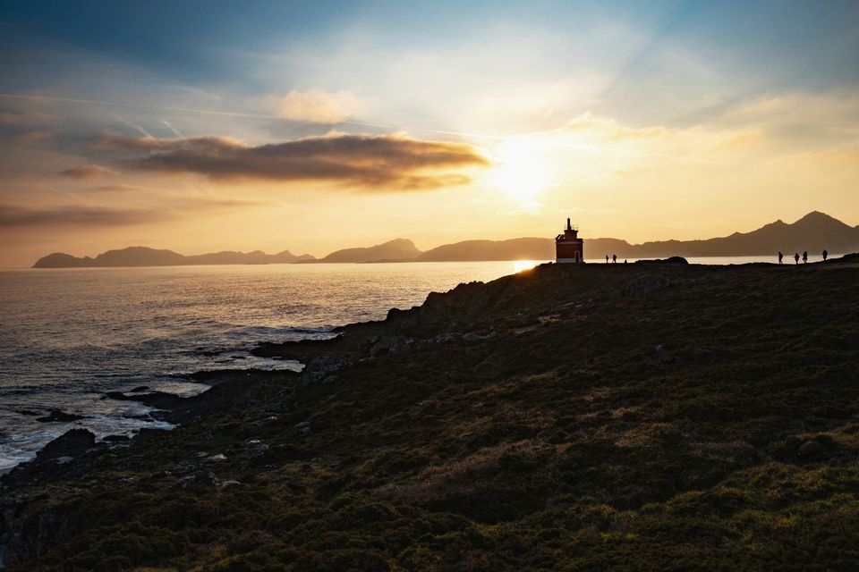 Sagome di un gruppo WeRoad che ammira il tramonto accanto a un faro rosso su una scogliera erbosa a picco sull'oceano