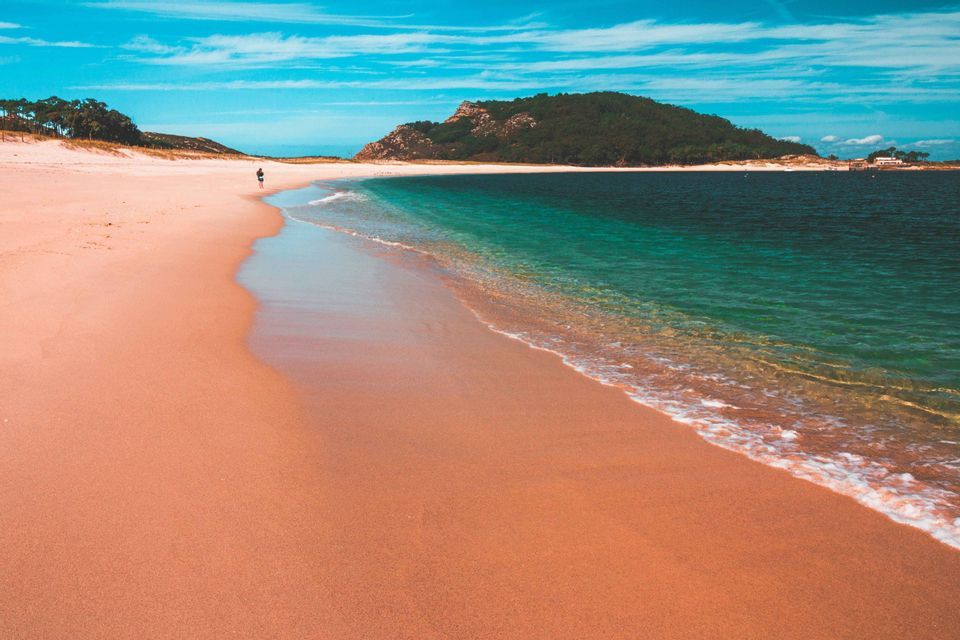 Una spiaggia ampia e sinuosa con sabbia rosata incontra il mare limpido e turchese sotto un cielo azzurro, con una persona che cammina in lontananza.