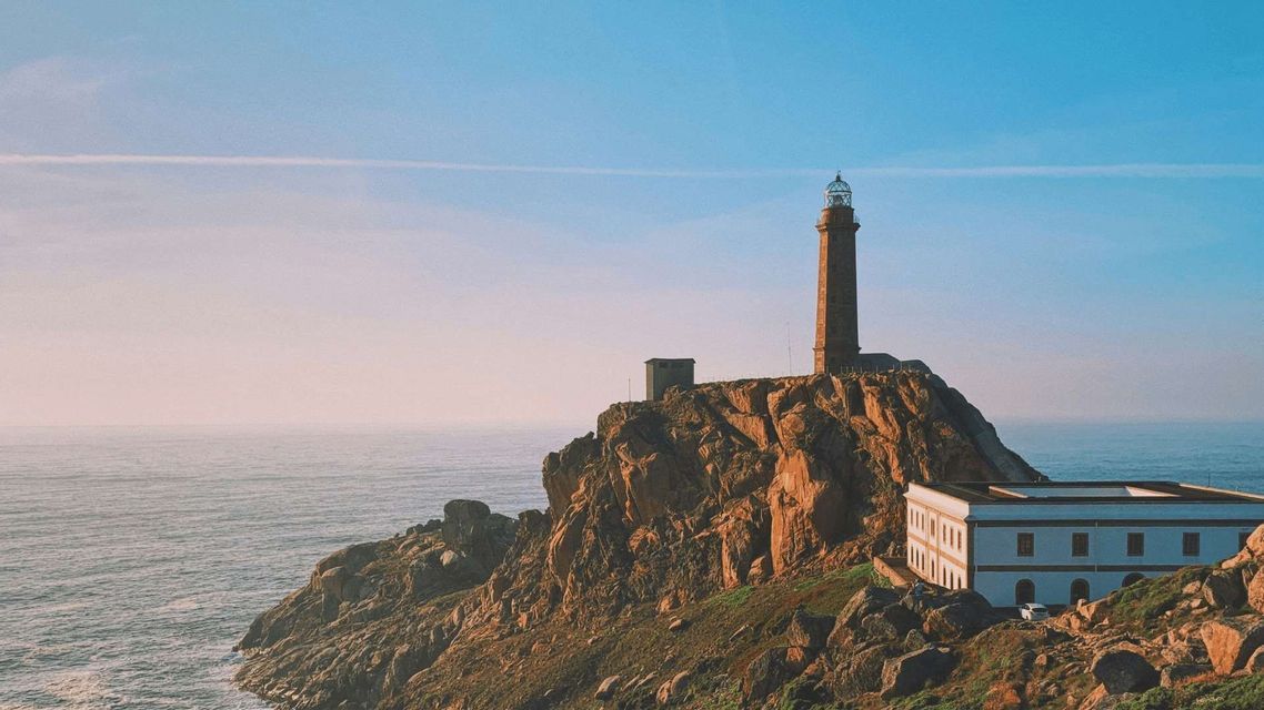 Un faro di pietra si erge su una scogliera aspra e rocciosa che domina un mare calmo sotto un cielo azzurro e limpido.