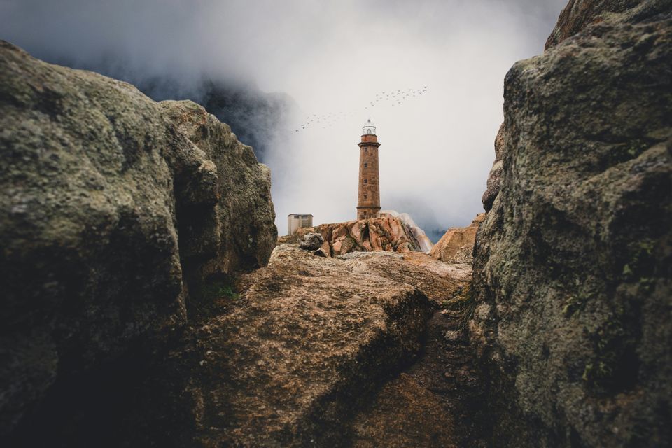 Un alto faro in pietra su una costa rocciosa, visto tra due grandi massi mentre gli uccelli volano in un cielo nebbioso.
