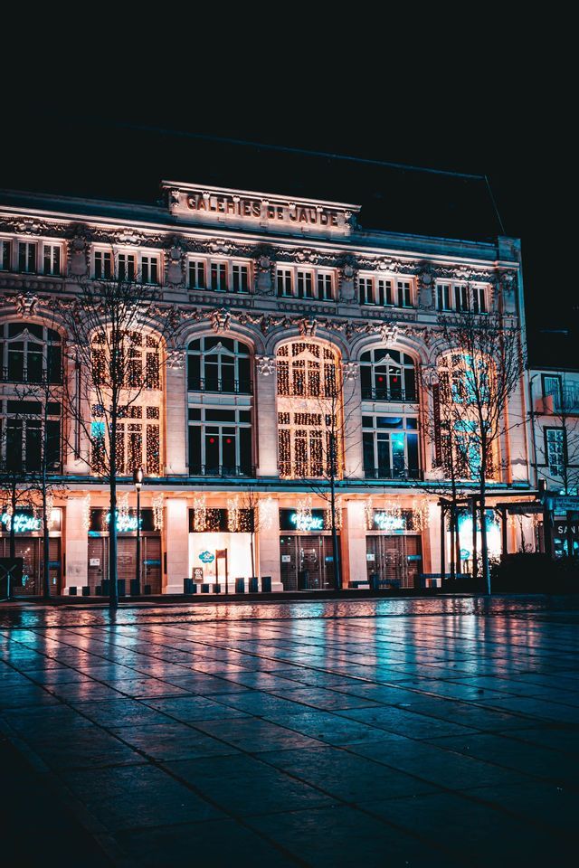 Le bâtiment orné des Galeries de Jaude illuminé la nuit, ses lumières se reflétant sur la rue pavée mouillée.