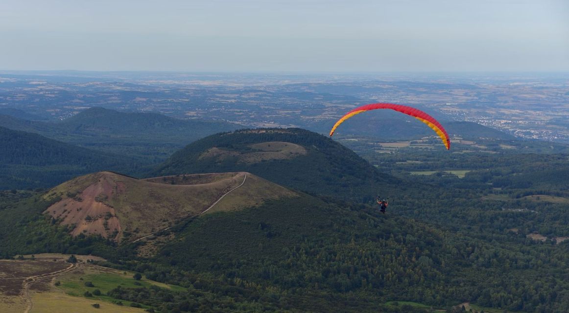 Une personne faisant du parapente avec une aile rouge et jaune au-dessus de collines verdoyantes et d'un cratère volcanique.