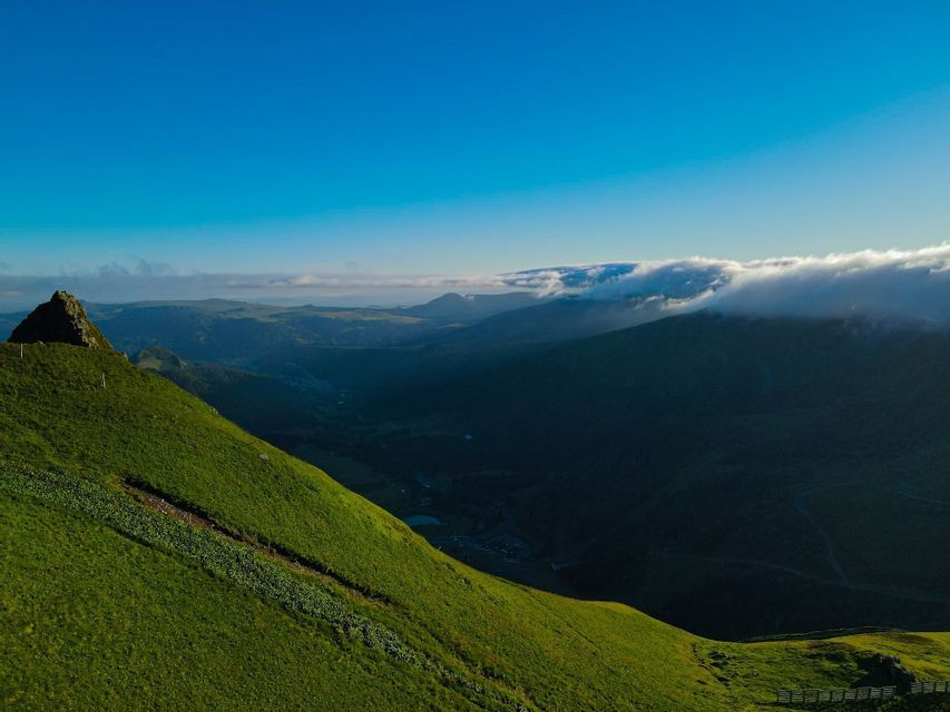 Une montagne verdoyante et escarpée domine une vallée ombragée, avec des sommets lointains partiellement couverts par les nuages sous un ciel bleu clair.