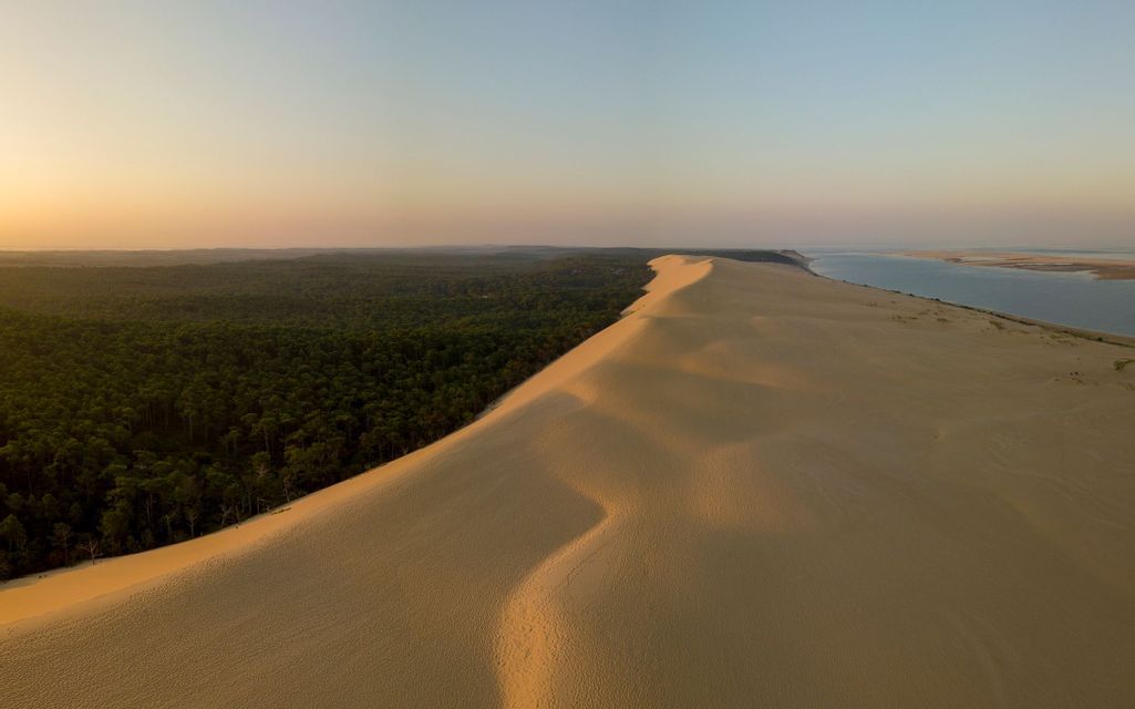 Une vaste dune de sable s'étend le long d'une forêt dense et d'un plan d'eau au coucher du soleil.