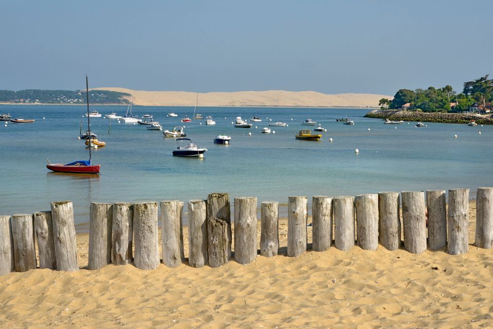 Des bateaux sont amarrés dans une baie paisible, face à une grande dune de sable, le tout vu depuis une plage de sable bordée d'une clôture en bois.