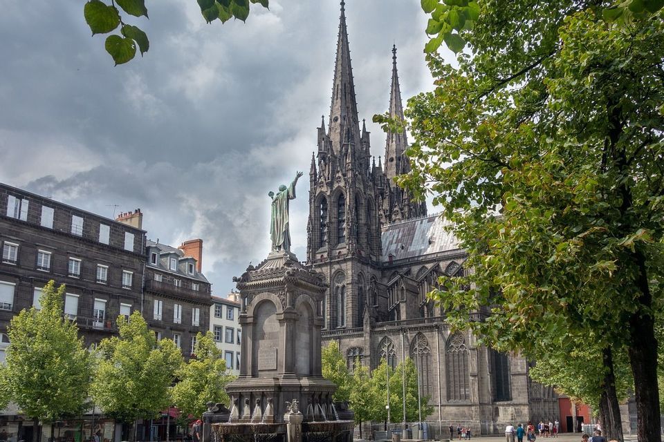 Une sombre cathédrale gothique aux doubles flèches domine une place de ville ornée d'une statue proéminente et d'une fontaine.