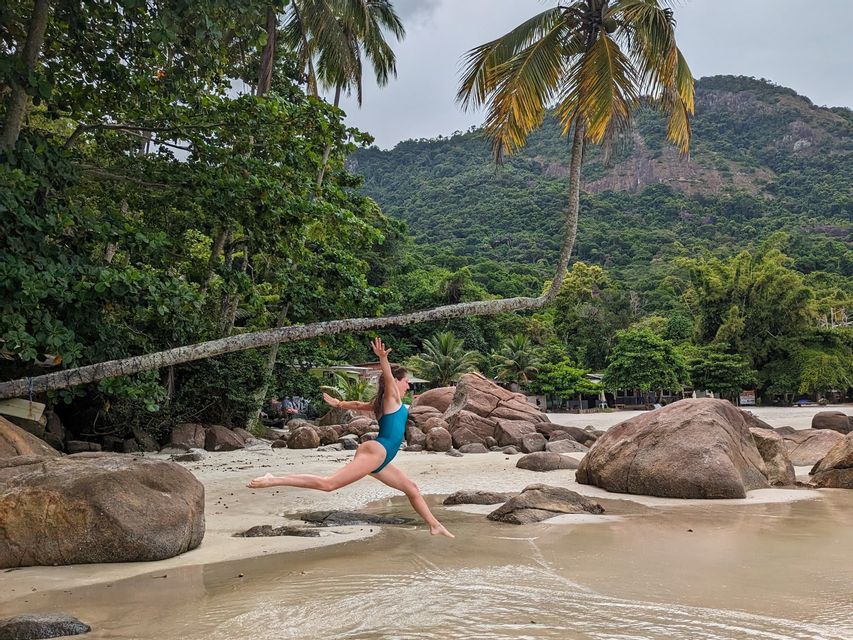 Una mujer en traje de baño azul salta en el aire en una playa de arena con grandes rocas, palmeras y una montaña boscosa de fondo.