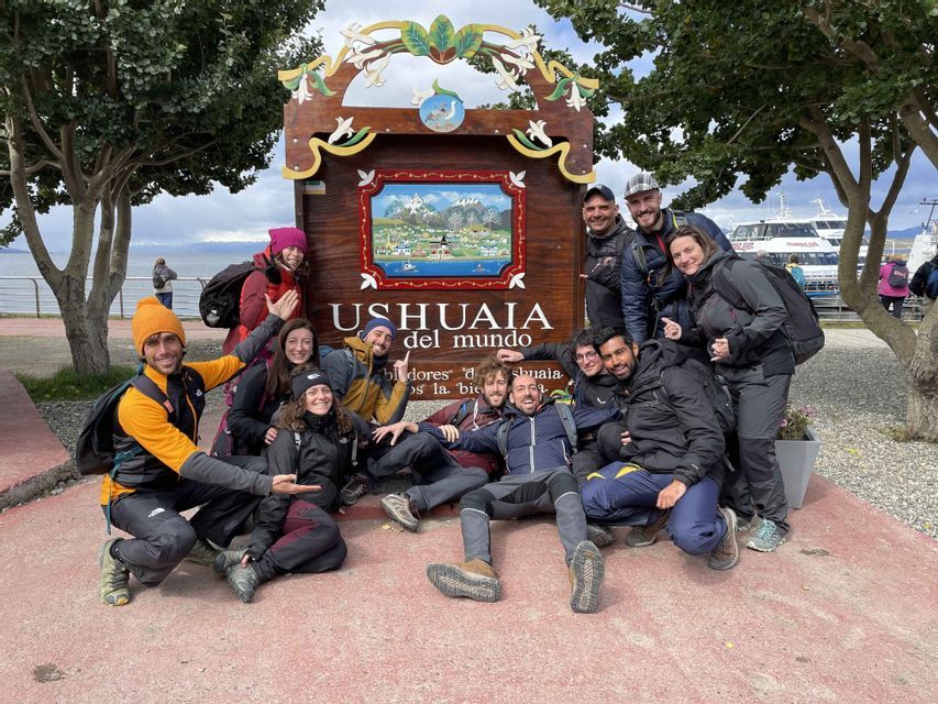 Eine WeRoad-Gruppe posiert lächelnd am Ufer vor dem großen Holzschild 'Ushuaia del mundo'.