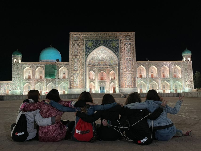 A WeRoad group trip of six women sit on the ground with arms linked, facing an ornate, illuminated building at night.