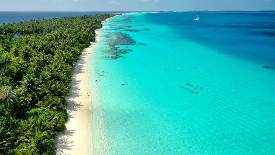 An aerial view of a long tropical island with a white sand beach, palm trees, and clear turquoise water with a sailboat in the distance.