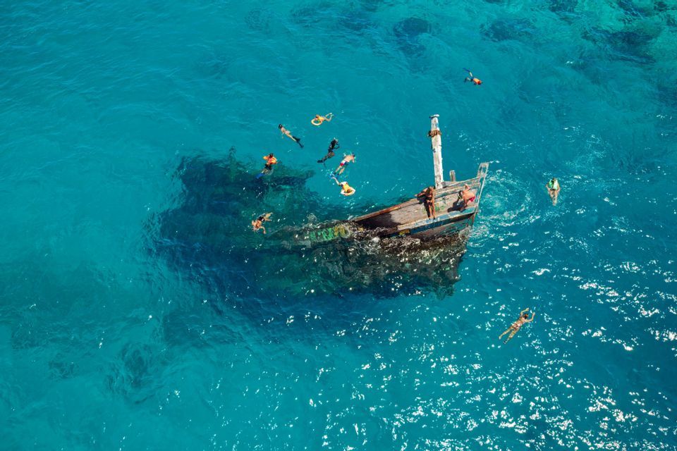 An aerial view of a WeRoad group trip snorkeling around a shipwreck in vibrant turquoise water.