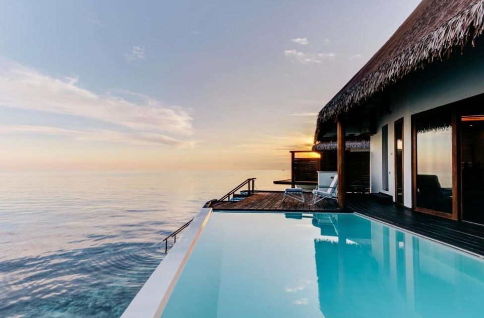 An overwater bungalow with a thatched roof and an infinity pool sits above the ocean at sunset.