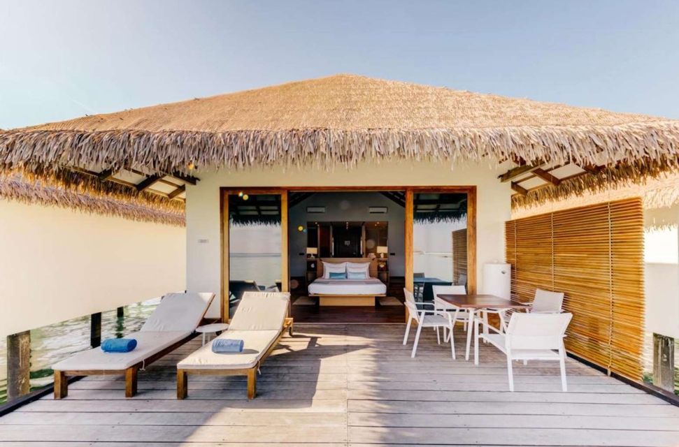 An overwater bungalow with a thatched roof features a wooden deck with lounge chairs and a table, with the bedroom visible inside.
