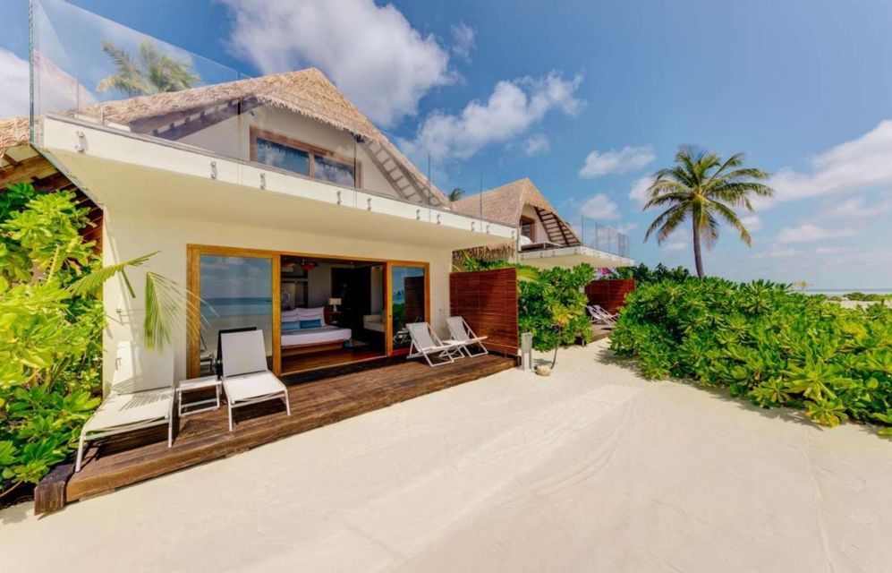A beachfront villa with a thatched roof and a wooden deck with lounge chairs, situated on a white sand beach next to tropical plants.