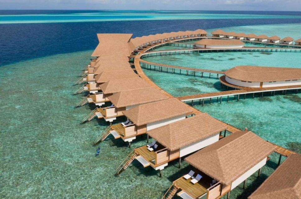 An aerial view of overwater bungalows with thatched roofs connected by walkways over a turquoise sea.