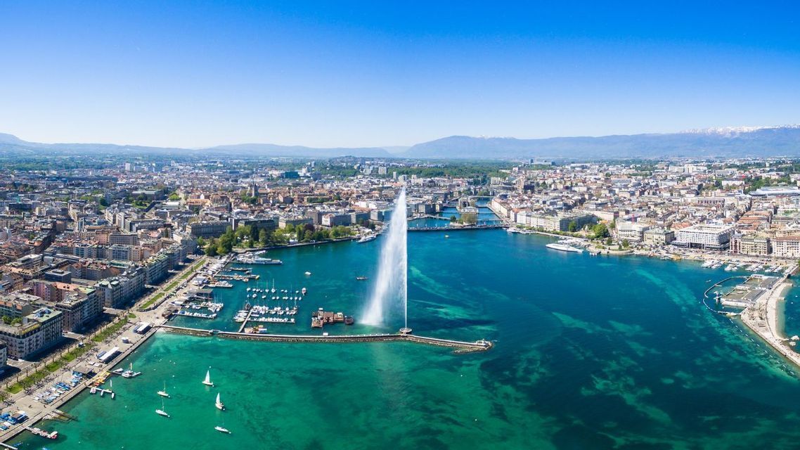 Una vista aérea de una gran fuente brotando agua desde una bahía turquesa, con una densa ciudad y montañas distantes.