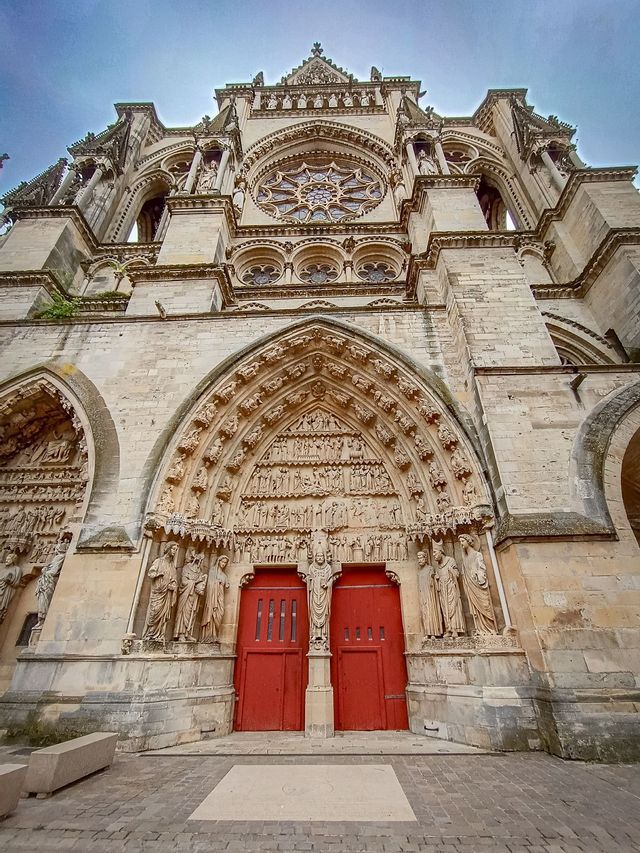 Una vista dal basso della facciata in pietra ornata di una cattedrale gotica con intagli intricati, un rosone e due porte rosse brillanti.