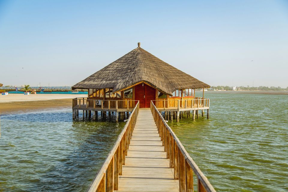 Un largo muelle de madera se extiende sobre el agua hacia una cabaña única sobre pilotes con techo de paja, con una orilla arenosa al fondo.