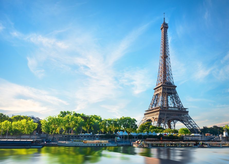 Der Eiffelturm ragt über der Seine empor, mit Booten, die am von Bäumen gesäumten Ufer unter einem klaren blauen Himmel festgemacht sind.