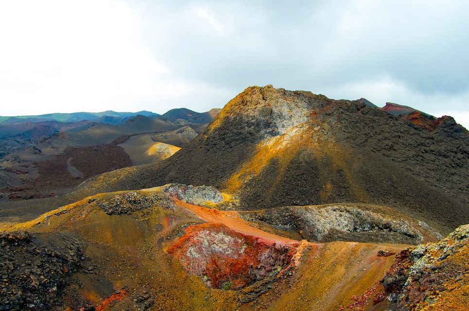 Un paisaje volcánico colorido con cráteres y colinas rocosas oscuras bajo un cielo nublado.