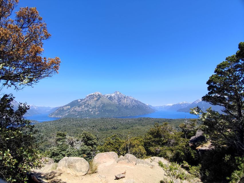 Una vista panoramica di un grande lago blu e di una catena montuosa, circondati da una fitta foresta, visti da un punto di osservazione elevato.