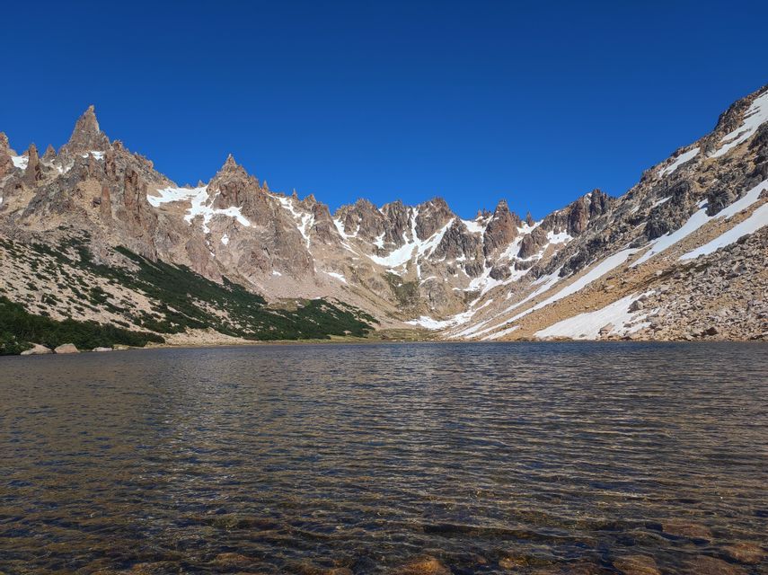 Un limpido lago di montagna riflette una catena montuosa frastagliata e innevata sotto un cielo azzurro senza nuvole.