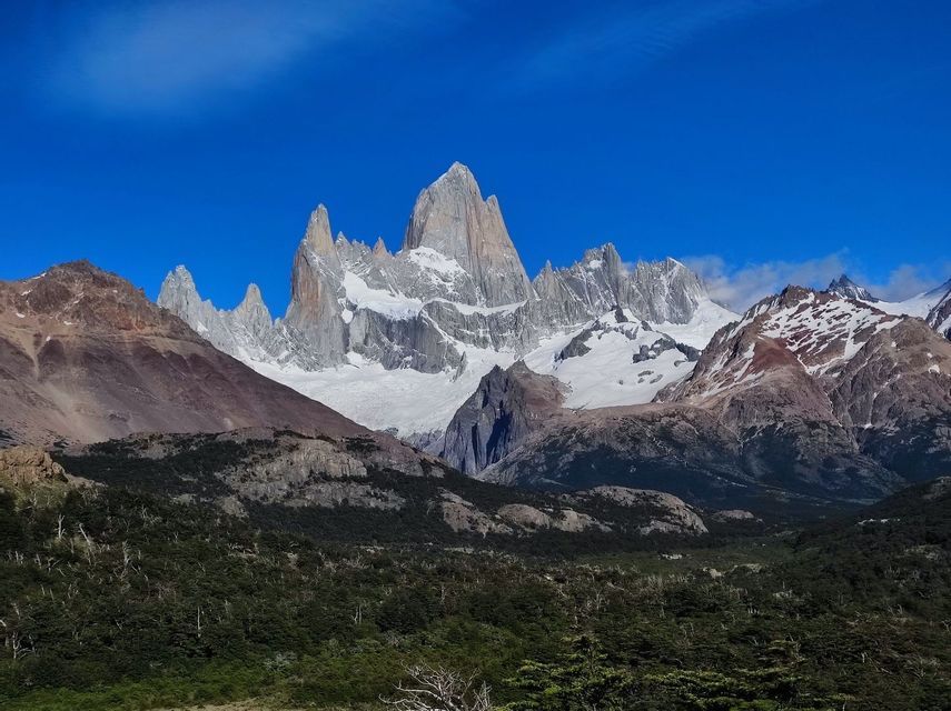 Montagne frastagliate e innevate si ergono sopra una verde valle boschiva, sotto un cielo azzurro e limpido.