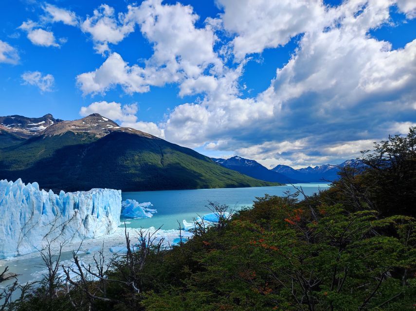 Un'immensa parete glaciale incontra un lago turchese pieno di iceberg, con montagne verdi sullo sfondo sotto un cielo azzurro nuvoloso.