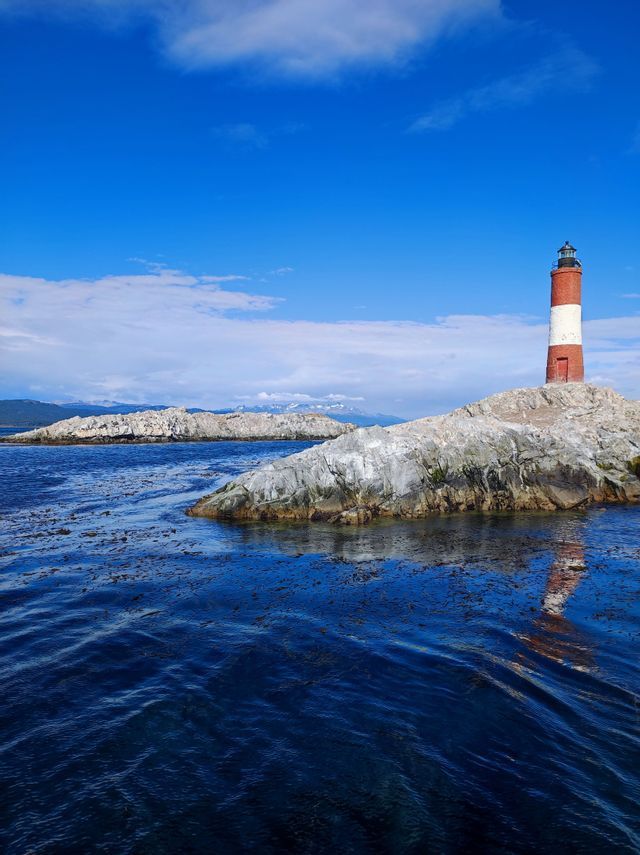 Un faro a strisce rosse e bianche si erge su un isolotto roccioso in mezzo all'acqua blu profonda sotto un cielo azzurro.