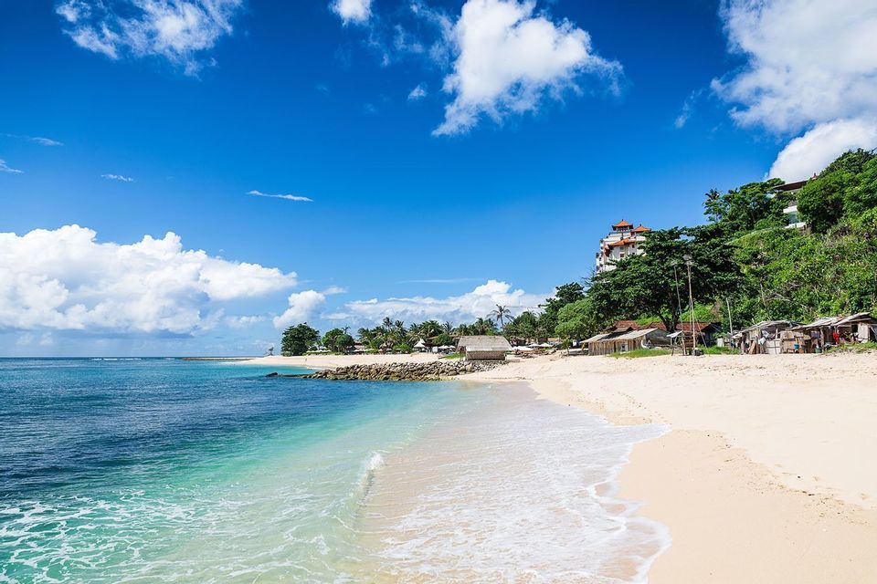 Playa tropical de arena con agua turquesa, palmeras y cabañas en la orilla bajo un cielo azul brillante con nubes blancas.