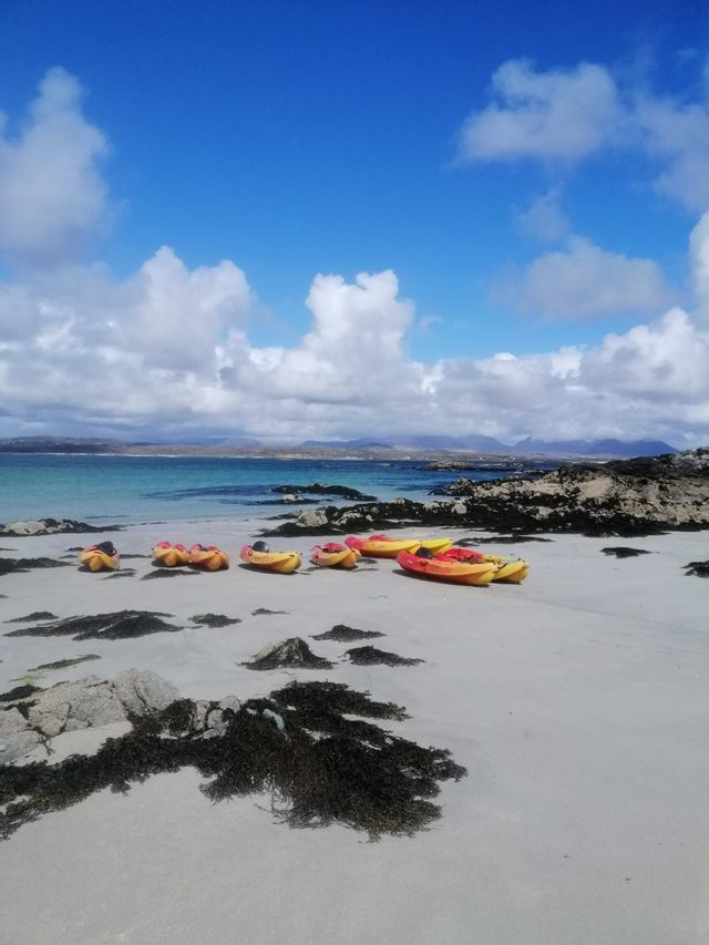 Diversi kayak colorati sono parcheggiati su una spiaggia di sabbia bianca con alghe scure, affacciata su una baia turchese sotto un cielo azzurro nuvoloso.