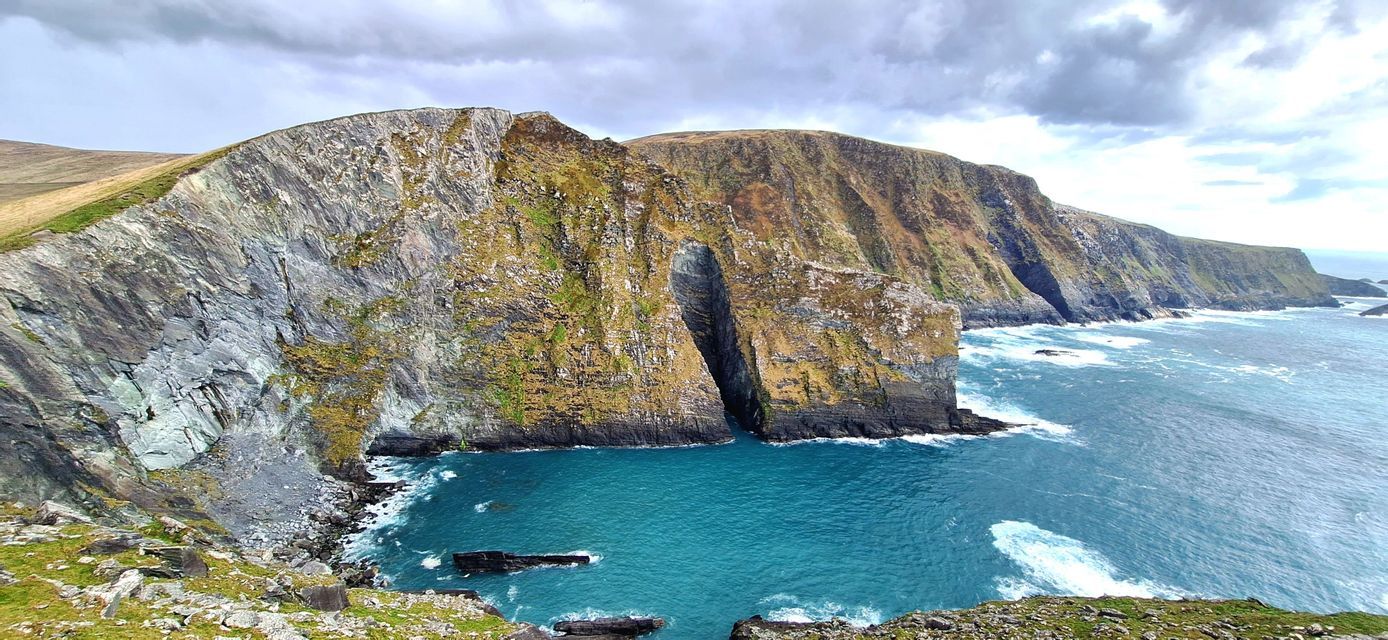 Ripide scogliere rocciose coperte di vegetazione verde e marrone scendono in una baia color turchese sotto un cielo nuvoloso.