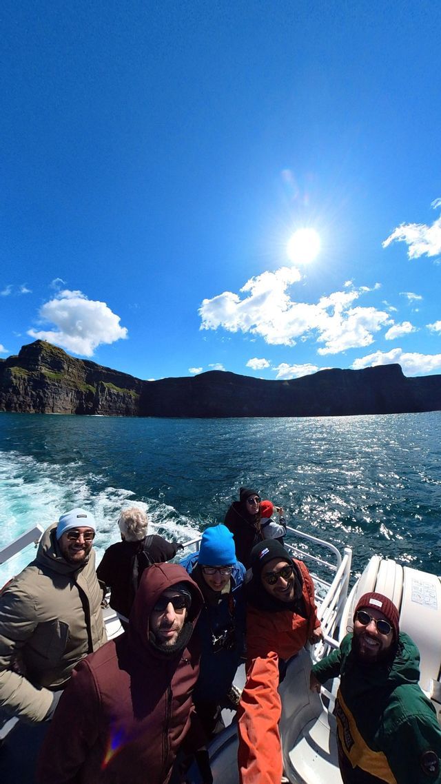 Un gruppo WeRoad si scatta un selfie su una barca che naviga oltre scogliere rocciose sotto un sole splendente e un cielo blu.
