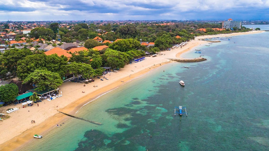 Vista aérea de una playa de arena curva que se une a aguas turquesas cristalinas, con exuberantes árboles y edificios costeros bajo un cielo nublado.