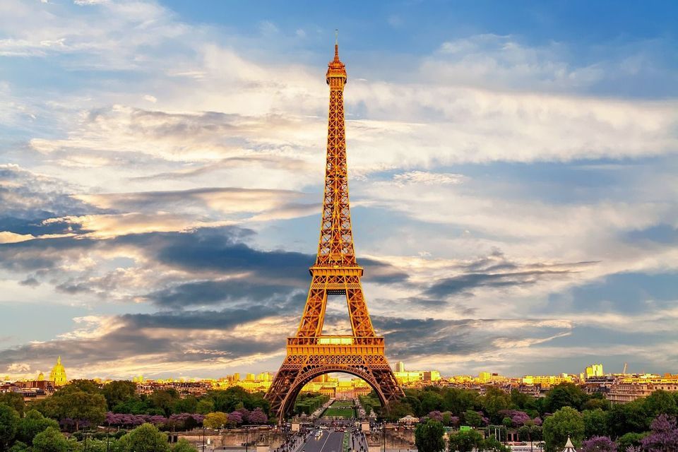 The Eiffel Tower stands tall against a partly cloudy sky at sunset, with the Paris cityscape and green trees below.