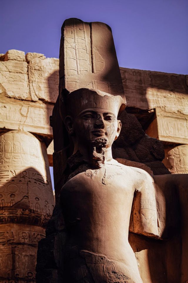 Una statua di pietra logorata dal tempo di un faraone si erge davanti ad antiche rovine sotto un cielo azzurro.