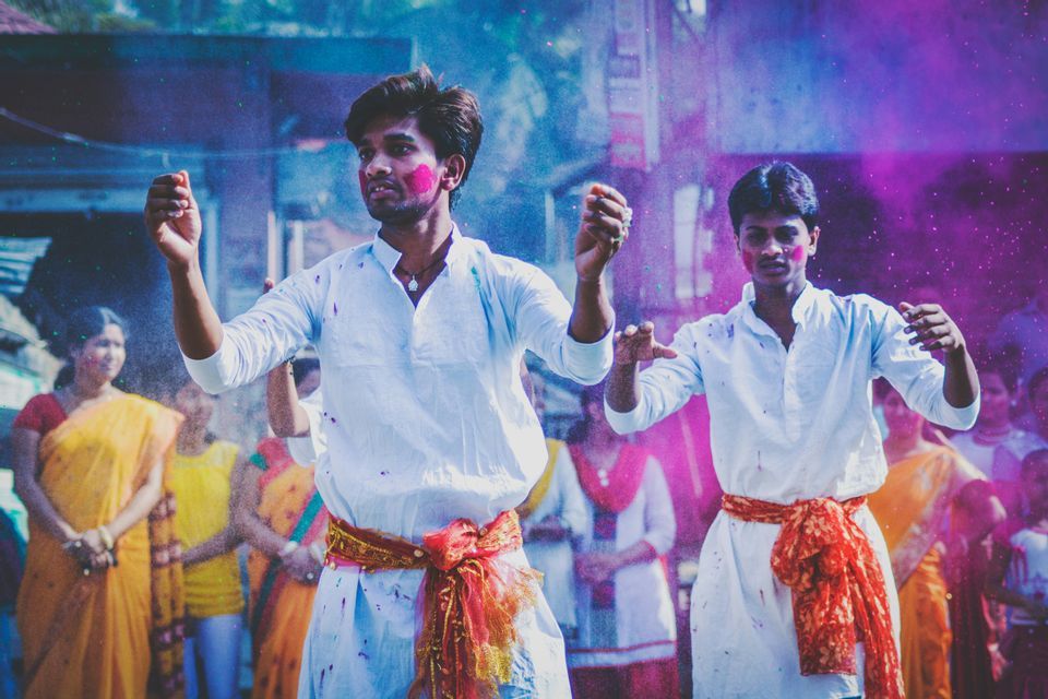 Dos hombres con trajes blancos y polvo de colores en sus caras bailan en medio de una nube de polvo morado.