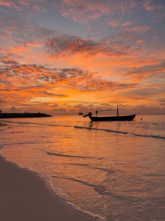Una barca in controluce galleggia sul mare vicino a una spiaggia sabbiosa sotto un cielo arancione intenso al tramonto.