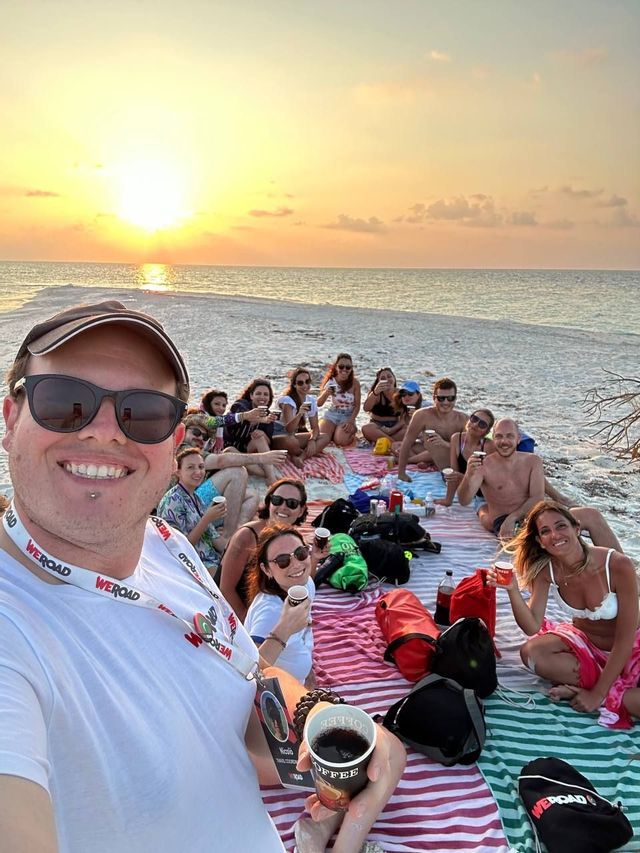 Un viaje en grupo de WeRoad se hace un selfie mientras hace un picnic en una playa de arena al atardecer.