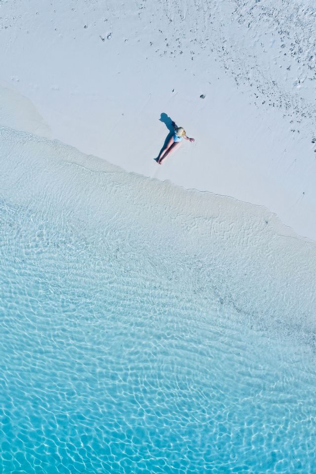 Top-down aerial view of a person in a swimsuit sunbathing on a white sand beach by clear turquoise water.