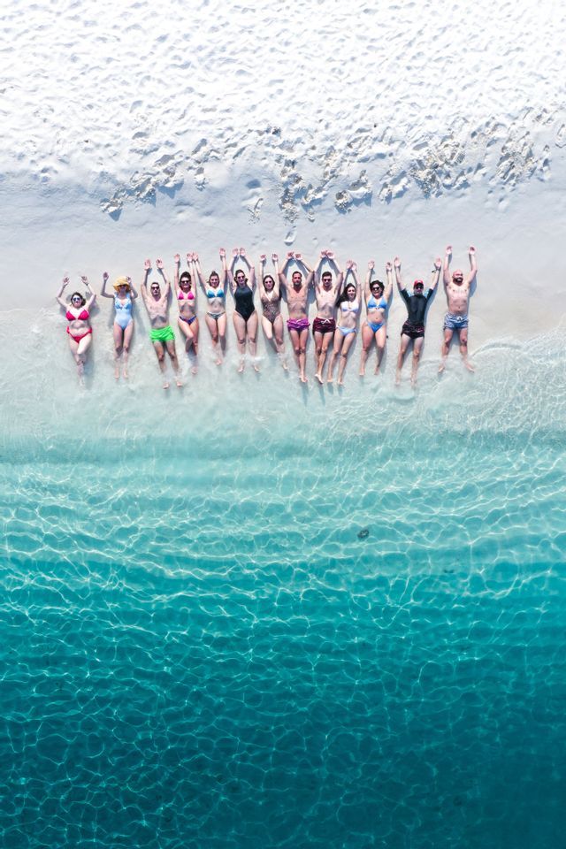Vista aerea di un gruppo WeRoad in viaggio, sdraiato in fila su una spiaggia di sabbia bianca con le braccia alzate e i piedi nell'acqua limpida e turchese.