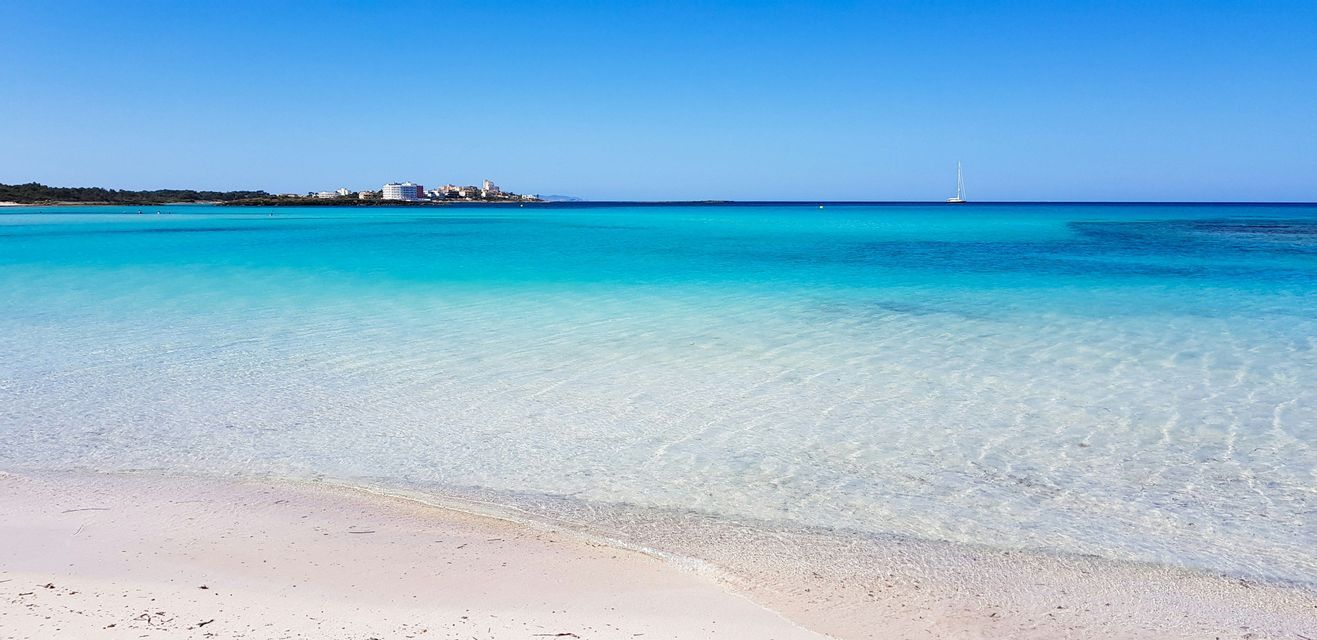 A view from a white sand beach across calm, turquoise water towards a distant coastline with buildings and a sailboat.